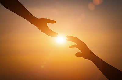 Two people's hands reaching to one another with sunset in the background.
